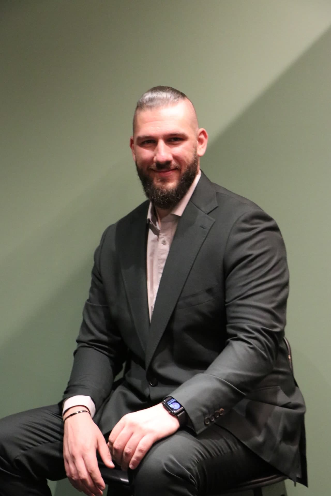 Daniel in a charcoal suit, seated against a green wall.